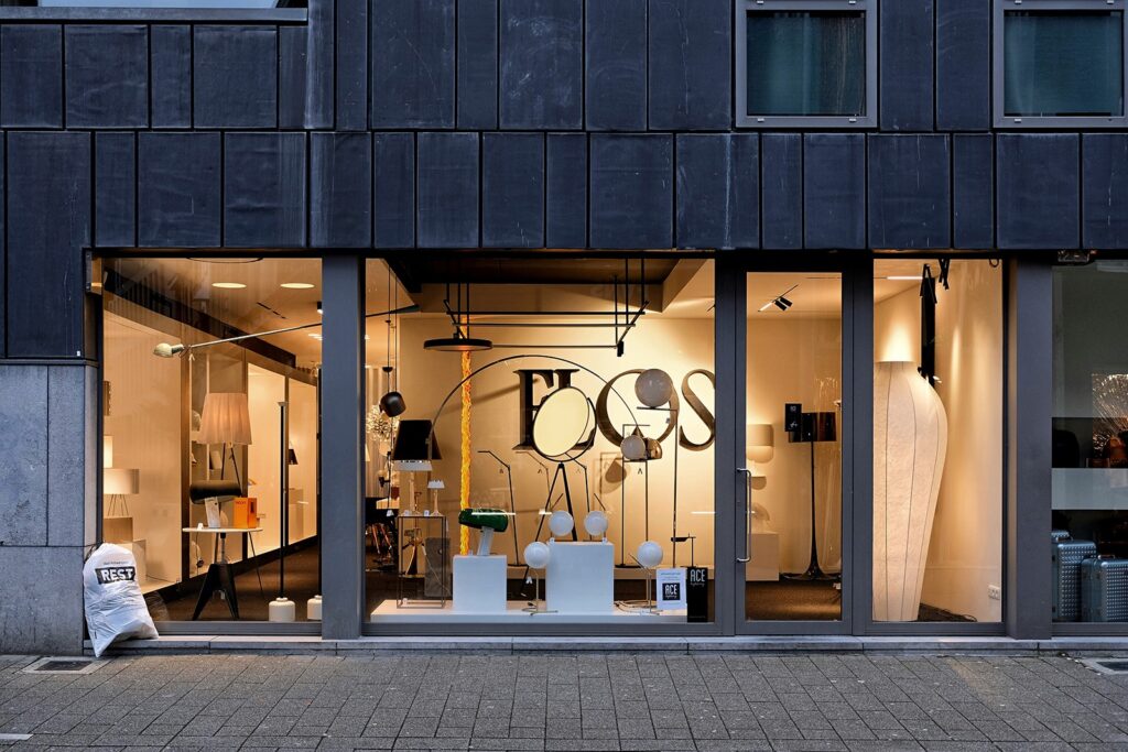 Storefront displaying various modern lamps and lighting fixtures against a sleek interior backdrop. Large glass windows reveal the showroom.