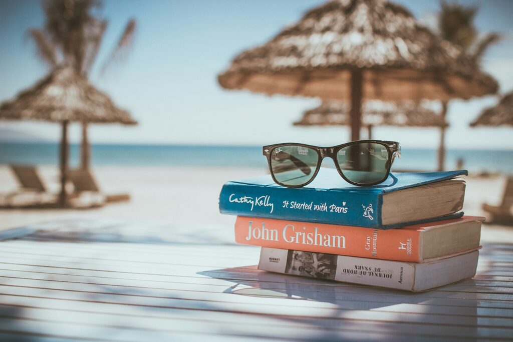 Stack of books topped with sunglasses on a table, with beach umbrellas and a clear sky in the background.
