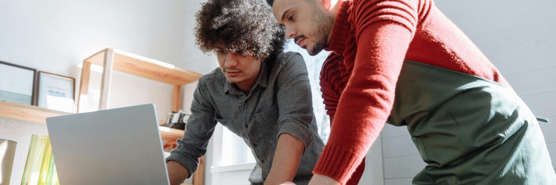 Two people stand in a kitchen, focused on a laptop screen. One wears a gray shirt, the other a red sweater and green apron.