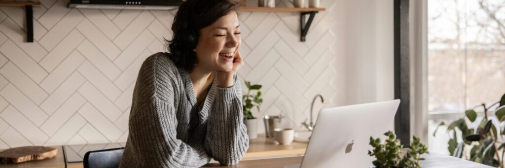 A person sits at a table, smiling while looking at a laptop. There are plants nearby and a white, tiled wall in the background.