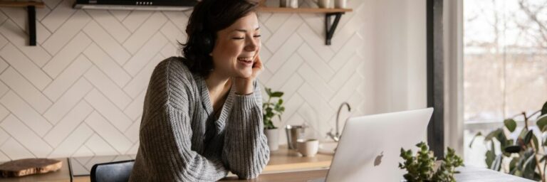 A person sits at a table, smiling while looking at a laptop. There are plants nearby and a white, tiled wall in the background.