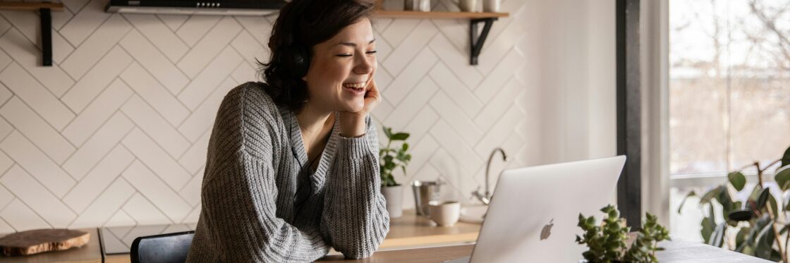 A person sits at a table, smiling while looking at a laptop. There are plants nearby and a white, tiled wall in the background.