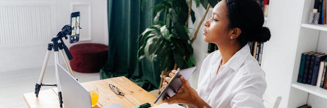 How to Optimize Your YouTube Channel A woman sits at a desk with a tablet, a smartphone on a tripod, a laptop, and a notebook. She appears to be recording or streaming in a home office setting.