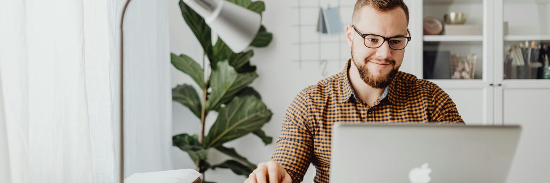 How to make a video A Beginner’s Guide A person with glasses and a beard is sitting at a desk, smiling while using a laptop. A lamp and plant are in the background.
