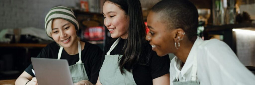 Three people wearing aprons smile while looking at a laptop in a casual setting.
