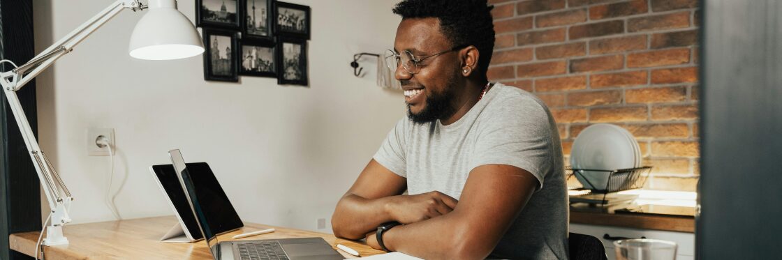 Man sitting at a wooden desk, looking at a laptop screen, smiling. There's a tablet and lamp on the desk, with framed photos on a brick wall in the background.
