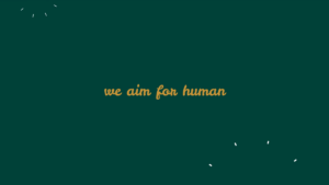 Text "we aim for human" in gold cursive font centered on a dark green background, with small white specks in the corners.