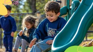 Young boy sliding down a playground slide with a smile, while two children sit and stand nearby on a sunny day.