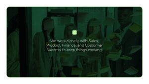 A group of four people stands in front of a glass wall covered with sticky notes, discussing work. Text overlay reads: "We work closely with Sales, Product, Finance, and Customer Success to keep things moving.