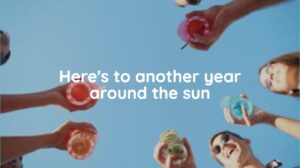 People holding colorful drinks in a circle, viewed from below against a blue sky, with the text "Here’s to another year around the sun" in the center.